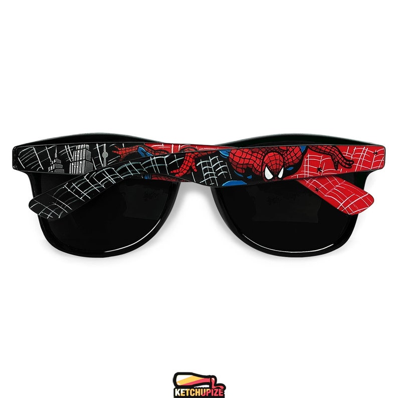 Picture of black sunglasses, hand-painted with a Spiderman comic theme design, showing Spiderman against spider web and the city skyline, in red, blue and black colors.