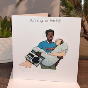 May include: White greeting card with the text "running up that hill". The card features an illustration of a man carrying a person, holding a boombox. The man wears a blue shirt with the word "WAVE".