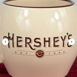 May include: Cream-colored Hershey's mug with a brown rim and handle. The Hershey's logo is prominently displayed in brown lettering, with the text "EST. 1894" below. The mug sits on a red surface.