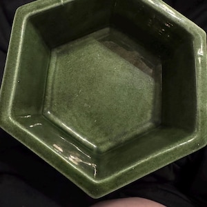 May include: A green, hexagonal ceramic planter with a glossy finish. The planter has a geometric design with angled sides and a deep interior. The color is a muted, earthy green, suitable for various plants and decor styles.