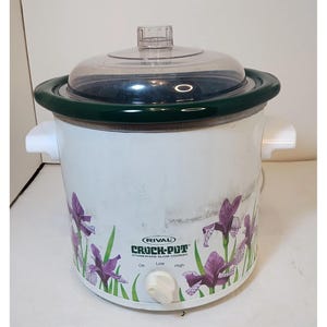 May include: White Rival Crock-Pot slow cooker with a clear lid and dark green trim. The slow cooker has purple iris flower designs and a control knob with settings for off, low, and high.