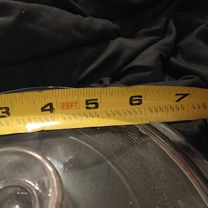 Könnte beinhalten: Ein klares Glasobjekt wird mit einem gelben Maßband gemessen. Das Maßband zeigt eine Messung von etwa 10 cm. Das Maßband ist mit schwarzen Zahlen und Brüchen markiert.