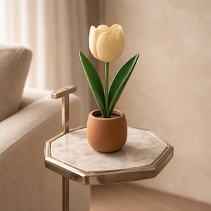 May include: A single, pale yellow tulip with green leaves in a small, terracotta-colored pot. The flower sits on a marble-topped, octagonal side table with a gold-colored metal frame. The table is next to a beige sofa.
