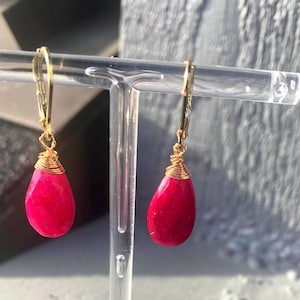 May include: A pair of teardrop-shaped earrings with a vibrant red hue. The gemstones are wrapped in gold-colored wire and suspended from gold-colored lever-back closures. The earrings are displayed on a clear acrylic stand.