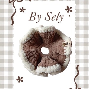 以下が含まれることがあります： 「By Sely」の文字が入った、茶色とクリーム色の手作りのシュシュ。柔らかく、質感のある素材で作られており、中央に穴が開いた円形です。縁はふわふわのクリーム色の素材でトリミングされています。