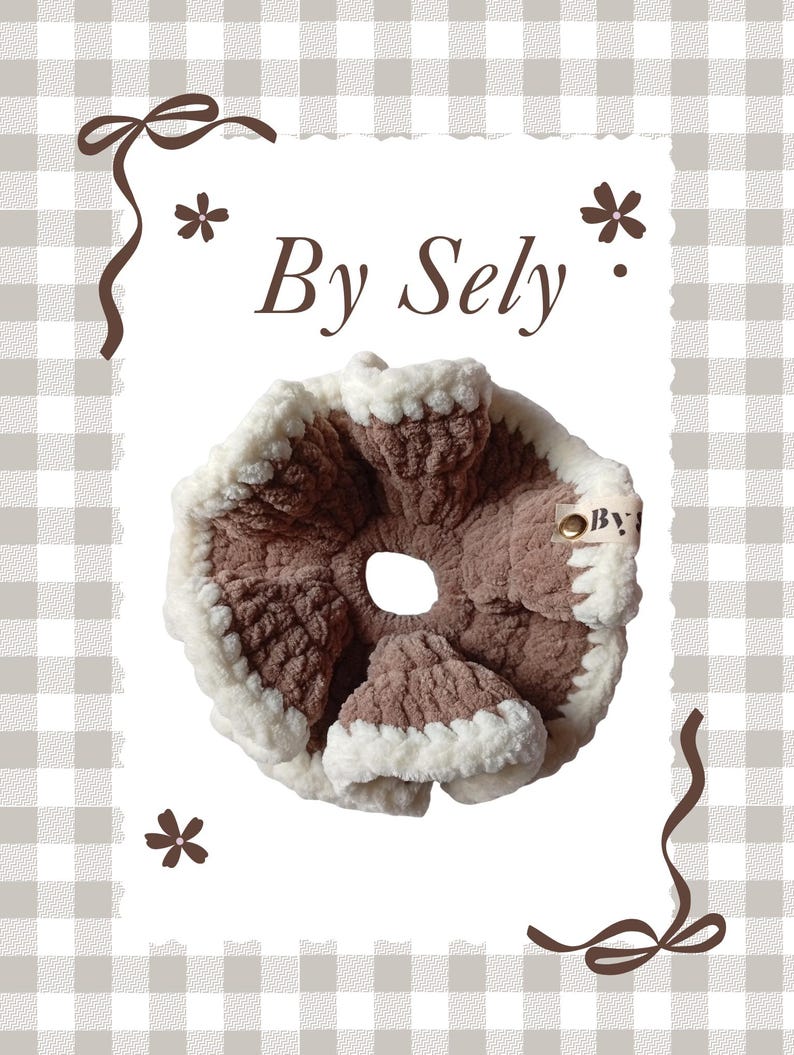 以下が含まれることがあります： 「By Sely」の文字が入った、茶色とクリーム色の手作りのシュシュ。柔らかく、質感のある素材で作られており、中央に穴が開いた円形です。縁はふわふわのクリーム色の素材でトリミングされています。