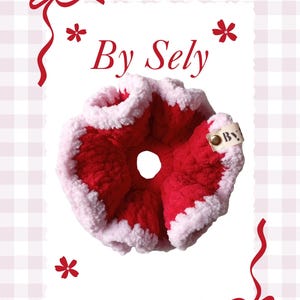 以下が含まれることがあります： 「By Sely」の文字が入ったタグが付いた、手作りの赤と白のシュシュ。 シュシュは柔らかく、質感のある素材で作られています。 装飾的な赤いリボンと花が、白い背景のシュシュを囲んでいます。