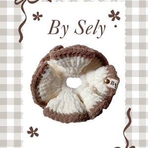 以下が含まれることがあります： クリーム色の中心と茶色の縁取りが特徴の手作りのシュシュ。シュシュには「By」と書かれた小さなタグが付いています。背景は「By Sely」の文字が入った白い長方形です。