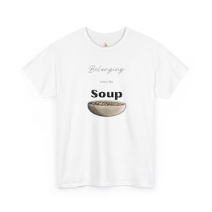 Può includere: T-shirt bianca con la scritta "Belonging tastes like Soup" in nero. Sotto, un'illustrazione di una ciotola di zuppa. Un capo casual per tutti i giorni.