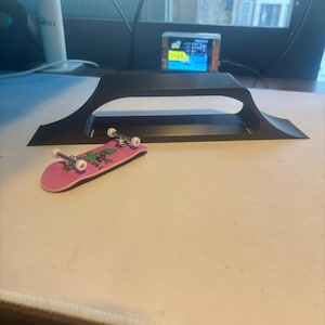 Puede incluir: Un monopatín rosa en miniatura con ruedas blancas y un gráfico verde, junto a una rampa negra. El monopatín tiene un gráfico verde que dice "Belly Button". La rampa es de plástico negro y está sobre una superficie blanca.