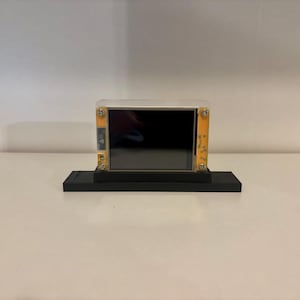 May include: A small electronic device with a black screen, encased in a clear plastic frame with yellow accents. It sits on a black rectangular base, suitable for display or integration into a project.