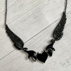 May include: Black necklace with a heart pendant and two wings on either side. The wings are detailed and have a gothic style.