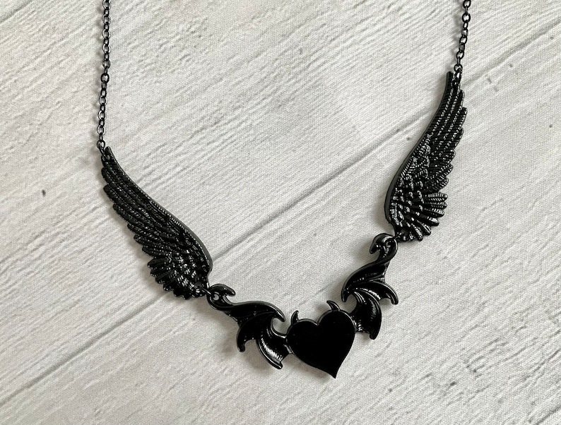 May include: Black necklace with a heart pendant and two wings on either side. The wings are detailed and have a gothic style.