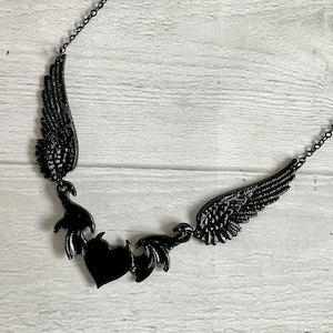 May include: Black necklace with a heart and two wings. The wings are made of metal and have a detailed design. The heart is also made of metal and has a black finish.