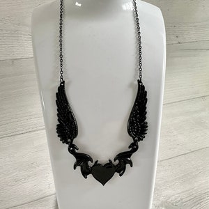 May include: A black necklace with a heart pendant and wings. The heart is surrounded by a decorative design that resembles bat wings.