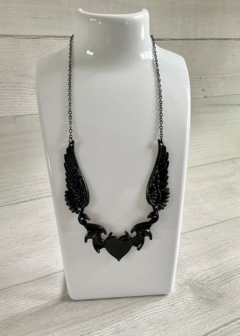 May include: A black necklace with a heart pendant and wings. The heart is surrounded by a decorative design that resembles bat wings.
