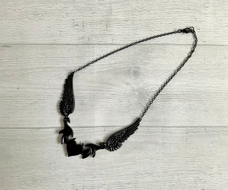 May include: A black necklace with a heart pendant and two wings on either side. The necklace is made of a delicate chain.