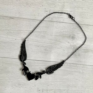 May include: A black necklace with a heart pendant and two wings on either side. The wings are detailed and intricate.