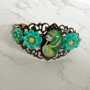 May include: A bronze bracelet with a green cameo featuring a woman with long black hair and a green dress. The bracelet is decorated with three turquoise flowers.