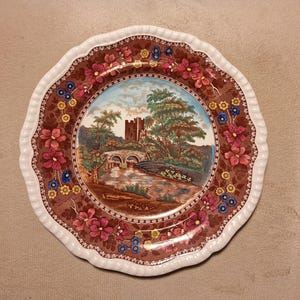 May include: A round ceramic plate with a scalloped white edge and a brown border decorated with pink, yellow, and blue flowers. The center features a colorful landscape illustration of a stone castle, a stone bridge, and a river with trees.
