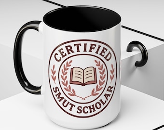 May include: A white ceramic mug with a black interior and handle. The mug features a brown circular design with the words "CERTIFIED SMUT SCHOLAR" around a book illustration.