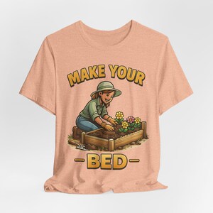 Könnte beinhalten: Pfirsichfarbenes T-Shirt mit einer Cartoon-Illustration einer Person, die gärtnert. Das Bild enthält den Text "MAKE YOUR -BED-" in Gelb mit Umrandung. Der Gärtner trägt ein grünes Hemd, blaue Jeans und einen Hut und kümmert sich um ein Blumenbeet.
