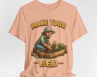 May include: Peach-colored t-shirt featuring a cartoon illustration of a person gardening. The image includes the text "MAKE YOUR -BED-" in a yellow, outlined font. The gardener is wearing a green shirt, blue jeans, and a hat, tending to a flower bed.