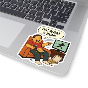 May include: A sticker with a cartoon illustration of a person laughing, holding a phone and a drink, and pointing at a TV showing a running figure. Text in a speech bubble reads "HA! WHAT A BUM!" A pizza box is on the floor. The sticker is on a laptop.