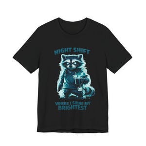 May include: Black t-shirt featuring a raccoon in scrubs holding a coffee mug. The raccoon illustration is in shades of blue and white, with the text "NIGHT SHIFT WHERE I SHINE MY BRIGHTEST" in blue.
