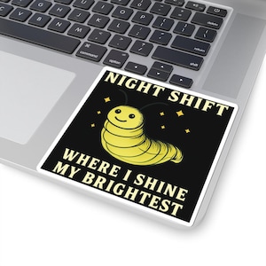 May include: A square sticker with the text "NIGHT SHIFT WHERE I SHINE MY BRIGHTEST" and a cartoon caterpillar. The caterpillar is yellow with a smiling face and is surrounded by small yellow stars. The sticker is on a laptop.