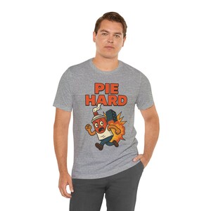 Könnte beinhalten: Graues T-Shirt mit dem Text "PIE HARD" in orangefarbenen Buchstaben. Das Shirt zeigt eine Cartoon-Grafik einer laufenden Kaffeetasse, die ein Gebäude hält, mit einem Explosionseffekt. Die Person trägt eine dunkelgraue Hose.