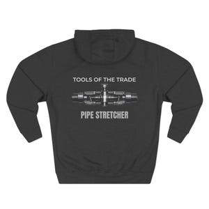 May include: Dark gray hoodie with a hood and long sleeves. The back of the hoodie features a graphic of a pipe stretcher tool with the text "TOOLS OF THE TRADE" above and "PIPE STRETCHER" below the graphic.