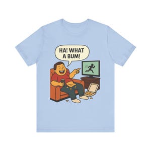 Puede incluir: Camiseta azul claro con una ilustración de dibujos animados. El diseño muestra a una persona riendo en un sofá, sosteniendo una bebida y viendo la televisión. El televisor muestra una figura corriendo, y el texto "HA! WHAT A BUM!" está en una burbuja.