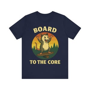 Puede incluir: Camiseta azul marino con un diseño de zarigüeya en monopatín. El diseño incluye un atardecer, pinos y el texto "BOARD TO THE CORE" en amarillo. Un diseño divertido e informal.