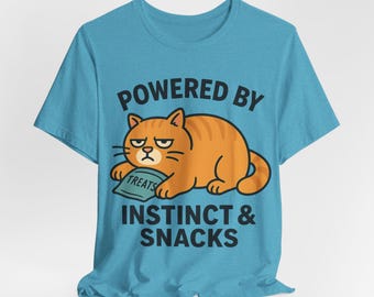 May include: A turquoise t-shirt with a cartoon image of an orange tabby cat. The cat has a grumpy face and is holding a bag of treats. The text on the shirt says "POWERED BY INSTINCT & SNACKS."