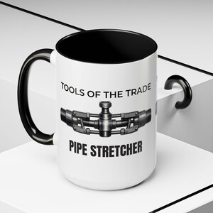 May include: A white ceramic mug with a black interior and handle. The mug features the text "TOOLS OF THE TRADE" above a detailed illustration of a pipe stretcher, with "PIPE STRETCHER" printed below. The mug is a humorous gift for a tradesperson.