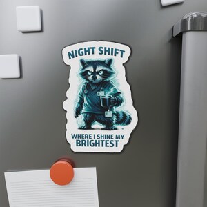 May include: A magnet featuring a raccoon in a teal medical uniform, holding a mug. The text "NIGHT SHIFT" is above the raccoon, and "WHERE I SHINE MY BRIGHTEST" is below. The raccoon has an intense expression.
