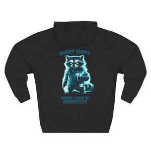 May include: Black hooded sweatshirt with a raccoon graphic. The raccoon is depicted in a medical uniform, holding a cup. The text "NIGHT SHIFT" and "WHERE I SHINE MY BRIGHTEST" is printed in blue on the sweatshirt.