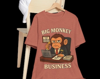 May include: A rust-colored t-shirt featuring a cartoon monkey in a suit, seated at a desk. The monkey is on the phone with a banana, with the text "BIG MONKEY BUSINESS" above and below the image. A white baseball cap rests on a wooden chair.