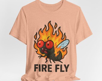 Può includere: T-shirt color pesca con una mosca dei cartoni animati in fiamme. La mosca ha grandi occhi rossi e un'espressione di panico. Le parole "FIRE FLY" sono stampate in lettere nere sotto.