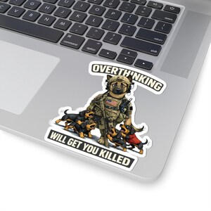 May include: A sticker showing a cartoon dog in military uniform with the text "OVERTHINKING WILL GET YOU KILLED." Several dachshund puppies surround the dog. The sticker is on a laptop.