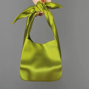 May include: A lime green satin shoulder bag with a knotted handle. The bag has a smooth, shiny texture and a simple, elegant design. The bag is held up against a gray background.