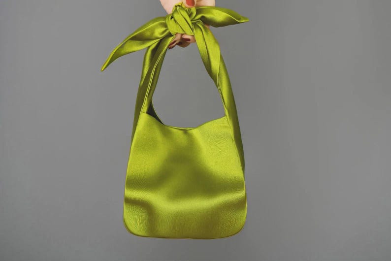 May include: A lime green satin shoulder bag with a knotted handle. The bag has a smooth, shiny texture and a simple, elegant design. The bag is held up against a gray background.