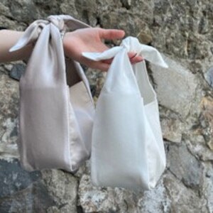 May include: Two small, rectangular handbags with a knotted strap. The handbag on the left is beige and the handbag on the right is white.
