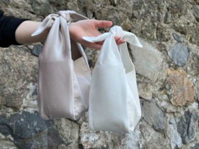 May include: Two small, rectangular handbags with a knotted strap. The handbag on the left is beige and the handbag on the right is white.