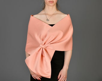 May include: A peach-colored satin shawl with a large bow detail. The shawl is draped over the shoulders and tied in the front, creating a voluminous effect. The garment is worn over a black top.