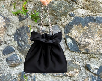 Peut inclure: Un sac à cordon en satin noir avec une bandoulière en chaîne argentée. Le sac a un haut plissé et une forme arrondie.