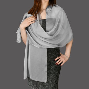 Puede incluir: Una bufanda gris claro y transparente drapeada sobre un vestido. La bufanda está envuelta alrededor de los hombros y los brazos, con un extremo colgando. El vestido es de color oscuro con una apariencia texturizada. El fondo es gris liso.