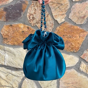 Puede incluir: Una bolsa de cordón de satén azul turquesa con una correa de cadena plateada. La bolsa tiene un borde con volantes y un diseño fruncido.