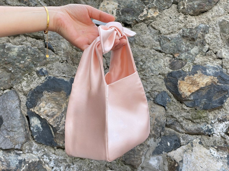 May include: A pale pink satin tote bag with a knotted handle. The bag is small and has a rectangular shape.